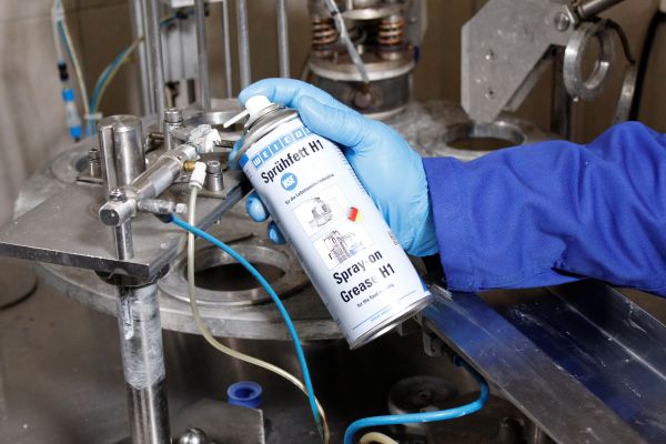 A person wearing a blue glove sprays WEICON Spray-on Grease H1 onto a metal fitting of an industrial machine.